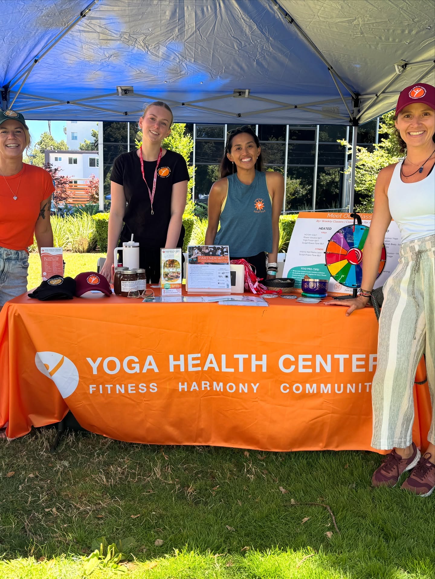 Four Yoga Health Center team members at the orange YHC booth under a blue canopy at Fit Fest